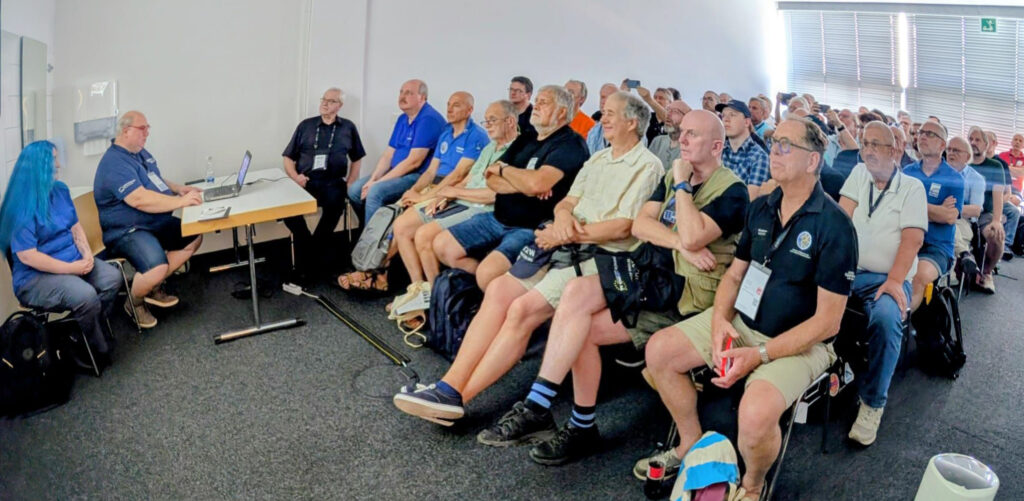 The futureGEO workshop at HAMRADIO 2026. At the desk in front, Peter Gülzow, president of AMSAT-DL, presents about the history and context of QO-100. Frank Zeppenfeldt was the second speaker, and defined the ESA's role with futureGEO, how Phase 1 would conclude, and what Phase 2 would entail. Michelle Thompson (far left) was the third and final speaker, and presented the working prototype from ORI for futureGEO submission. 